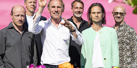 Auf dem Foto sind 6 Herren mittleren Alters zu sehen, sommerlich schick kekleidet. Der HIntergrund ist pink und drumherum bunte Blumen zu sehen