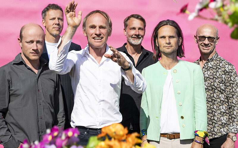 Auf dem Foto sind 6 Herren mittleren Alters zu sehen, sommerlich schick kekleidet. Der HIntergrund ist pink und drumherum bunte Blumen zu sehen