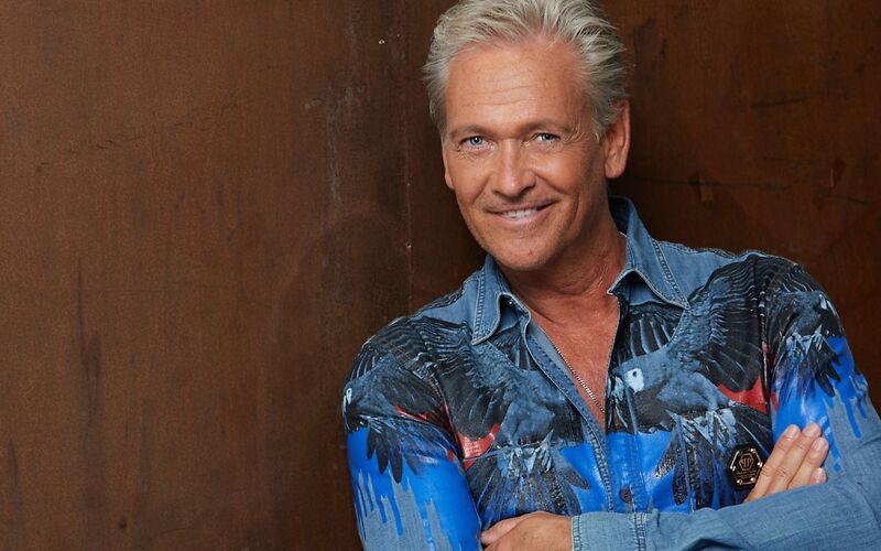 Mann grau melierte Haare, lächelt freundlich den Betrachter an, er lehnt mit den Armen verschränkt an einer Wand. ächelt