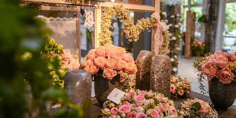 Auf dem Tisch in einem Ausstellungsraum stehen vier Vasen und zwei kleine Rosenkränze mit rosafarbigen Rosen. Im Hintergrund befindet sich ein Heidekranz in Form eines Herzens. Durch die Fenster sieht man die grüne Natur vom Rosengarten.