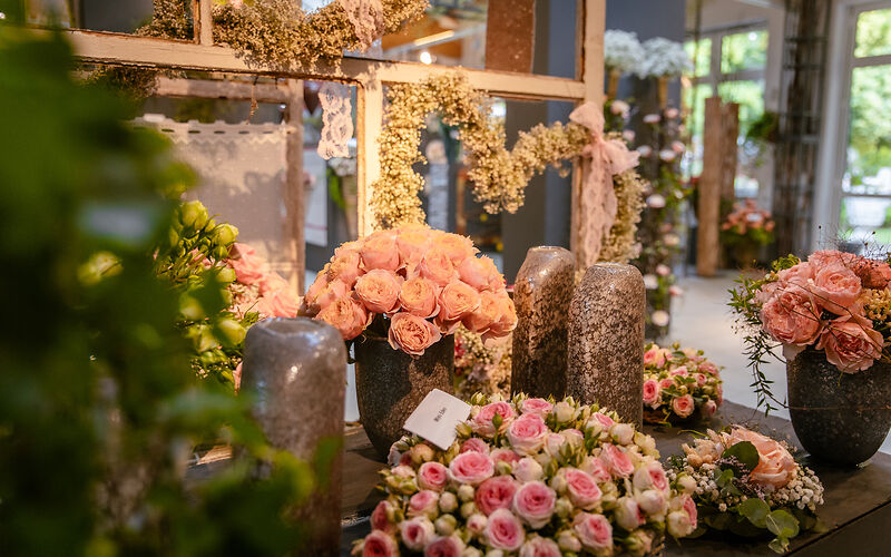 Auf dem Tisch in einem Ausstellungsraum stehen vier Vasen und zwei kleine Rosenkränze mit rosafarbigen Rosen. Im Hintergrund befindet sich ein Heidekranz in Form eines Herzens. Durch die Fenster sieht man die grüne Natur vom Rosengarten.