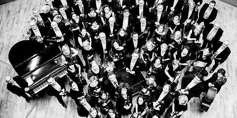 ein schwarz-weiß Bild zeigt ein Orchester der Zuschauer guckt von oben auf die Gruppe runter, die Muiker lächeln nach oben