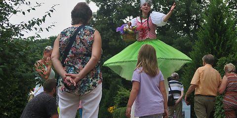 Eine bunt bekleidete Frau auf Stelzen begrüst spazierende Gäste im Park