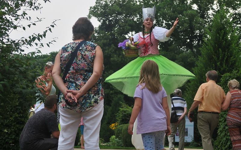 Eine bunt bekleidete Frau auf Stelzen begrüst spazierende Gäste im Park
