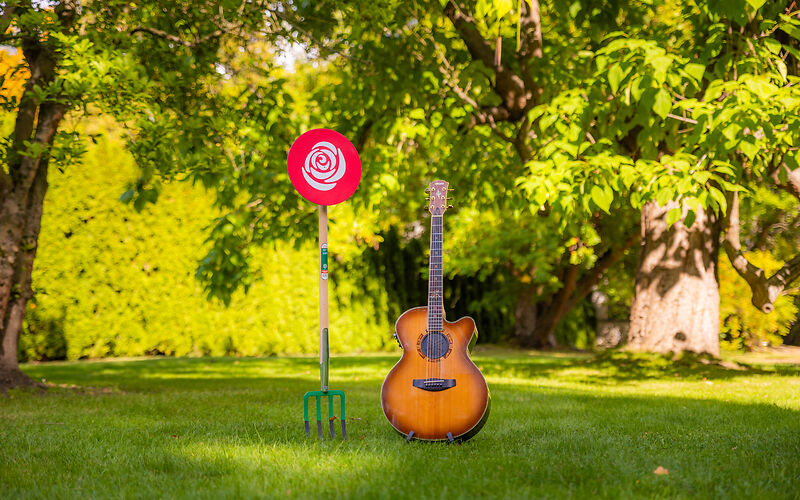 Auf einer Wiese unter mit Blättern bewachsenen Baumzweigen stehen eine Mistgabel, an welcher eine auf Papier gedruckte weiße Rose auf rotem Hintergrund oben befristigt ist, und eine Gitarre. 