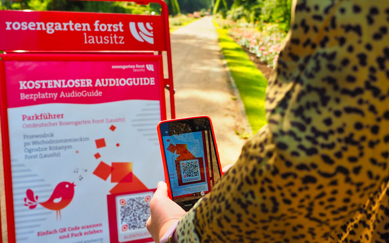 IM Vordergrund eine Person mit Handy, die einen QR Code vom Plakat scannt