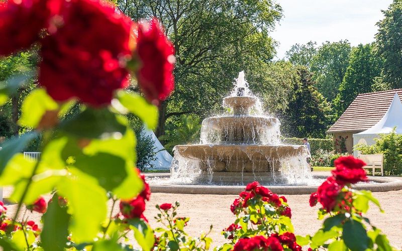 Dreistufiger Springbrunnen auf einer Sandfläche vor Bäumen mit roten Rosen im Vordergrund