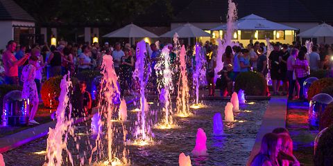 Die großen Wasserspiele in der Abenddämmerung, drumherum Menschen stehen und sitzend betrachten das bunte Schauspiel