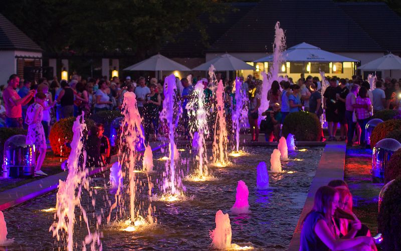 Die großen Wasserspiele in der Abenddämmerung, drumherum Menschen stehen und sitzend betrachten das bunte Schauspiel