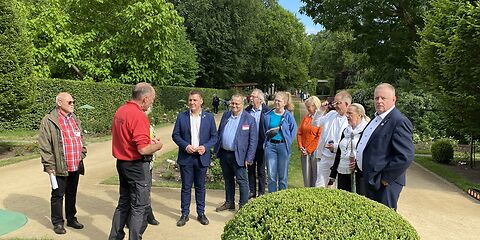 Die Gruppe steht vor länglich angelegten Rosenbeeten umgeben von Bäumen.