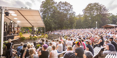 linke ist eine Open Air Bühne, davor eine Stufentribüne mit tausende sitzende Zuschauenden 