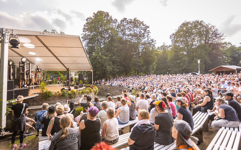 linke ist eine Open Air Bühne, davor eine Stufentribüne mit tausende sitzende Zuschauenden 
