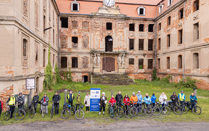 Die Gruppe steht mit ihren Fahrrädern vor der Ruine des Schlosses Brody. Das Gebäude ist u-förmig mit der offenen Seite nach vorn. Zum Haupteingang des mittleren Gebäudeteils führt eine breite mit Moss bewachsene Treppe. Neben der großen Tür sind links und rechts jeweils 2 Statuen, oben am Gebäude befindet sich eine Uhr.