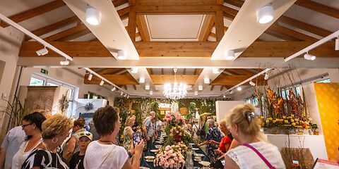 Die Halle mit der Schnittrosenschau, vom Eingang aus fotografiert. In der Mitte steht eine lange Tafel mit Sammeltassen an beiden Seiten und Blumenbuketts in der Mitte. Um den Tisch herum stehen Besucher der Schnittrosenschau.