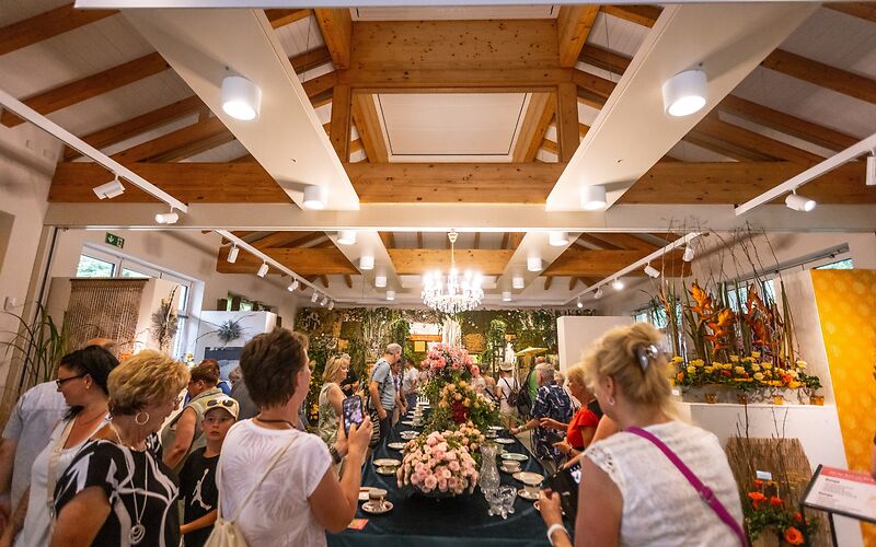 Die Halle mit der Schnittrosenschau, vom Eingang aus fotografiert. In der Mitte steht eine lange Tafel mit Sammeltassen an beiden Seiten und Blumenbuketts in der Mitte. Um den Tisch herum stehen Besucher der Schnittrosenschau.