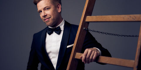 Tom Gaebel in einem schwarzen Smoking angelehnt an eine Leiter. 