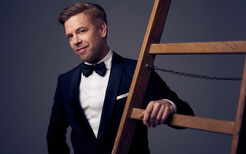 Tom Gaebel in einem schwarzen Smoking angelehnt an eine Leiter. 