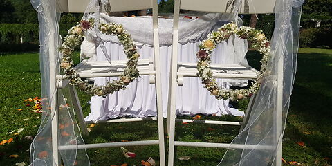 zwei weiße Stühle geschmückt mit Tüll und Blumenkränze, dahinter ein Tisch mit weißen Laken für die freie Trauung