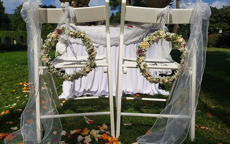 zwei weiße Stühle geschmückt mit Tüll und Blumenkränze, dahinter ein Tisch mit weißen Laken für die freie Trauung