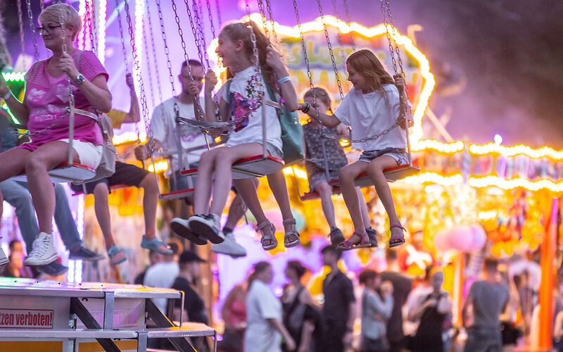 ein Stimmungsbild mit zeigt ein Kettenkarusell mit lachenden jungen Menschen im Hintergrund ein weiteres Fahrgeschäft beleuchtet