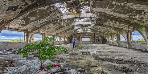 Das Foto zeigt eine sehr große, leere Fabrikhalle. Die ehemalige Fabrik wird heute als Gründer- und Traumfabrik in Forst (Lausitz) genutzt. Durch die vielen großen Fensteröffnungen ohne Scheiben fällt viel Licht in den Raum. Von dort aus ist der Himmel mit blauen Wolken zu sehen. In der Halle wächst ein kleiner Baum, der einen starken Kontrast zur industriellen Umgebung bildet.