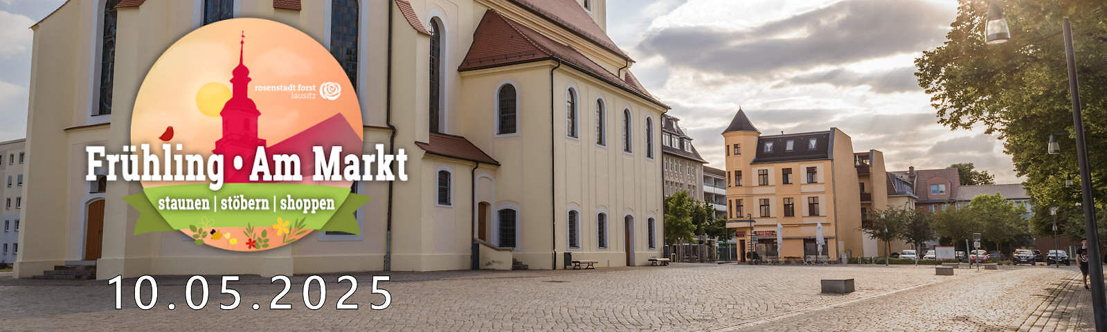 Links im Bild ist er untere Teil der Stadtkirche St. Nikolai mit dem leeren gepflasterten Marktplatz drum herum zu sehen. Im Hintergrund befinden sich Wohnhäuser und ein Parkplatz. Links ist ebenfalls des Logo "Frühling Am Markt" und das Datum 10.05.2025 zu sehen. 