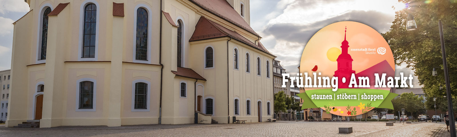 seitlicher Blick auf das Kirchenschiff der Stadtkirche (hellgelbes Gebäuden mit zweireihigen nach oben runden Fenstern) auf dem geplfasterten Marktplatz. Links befindet sich das Logo "Frühlung am Markt - staunen | stöbern | schoppen"
