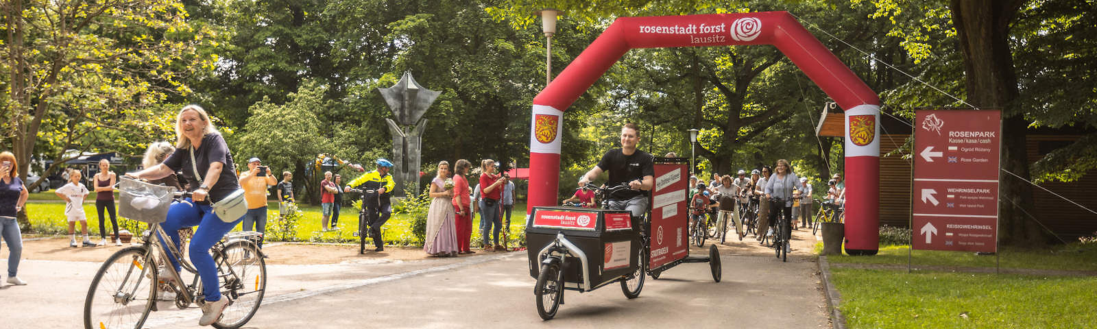 Auf einem Sandweg im Rosengarten ist ein roter Toorbogen mit der Aufschrift "stadt forst lausitz" aufgestellt. Auf dem Weg fahren die Bürgermeisterin Frau Taubenek und Herr Herrmann auf Fahrrädern. Weitere Fahrradfahren machen sich unter dem Bogen zum Start bereit. Rings herum stehen Zuschauer.